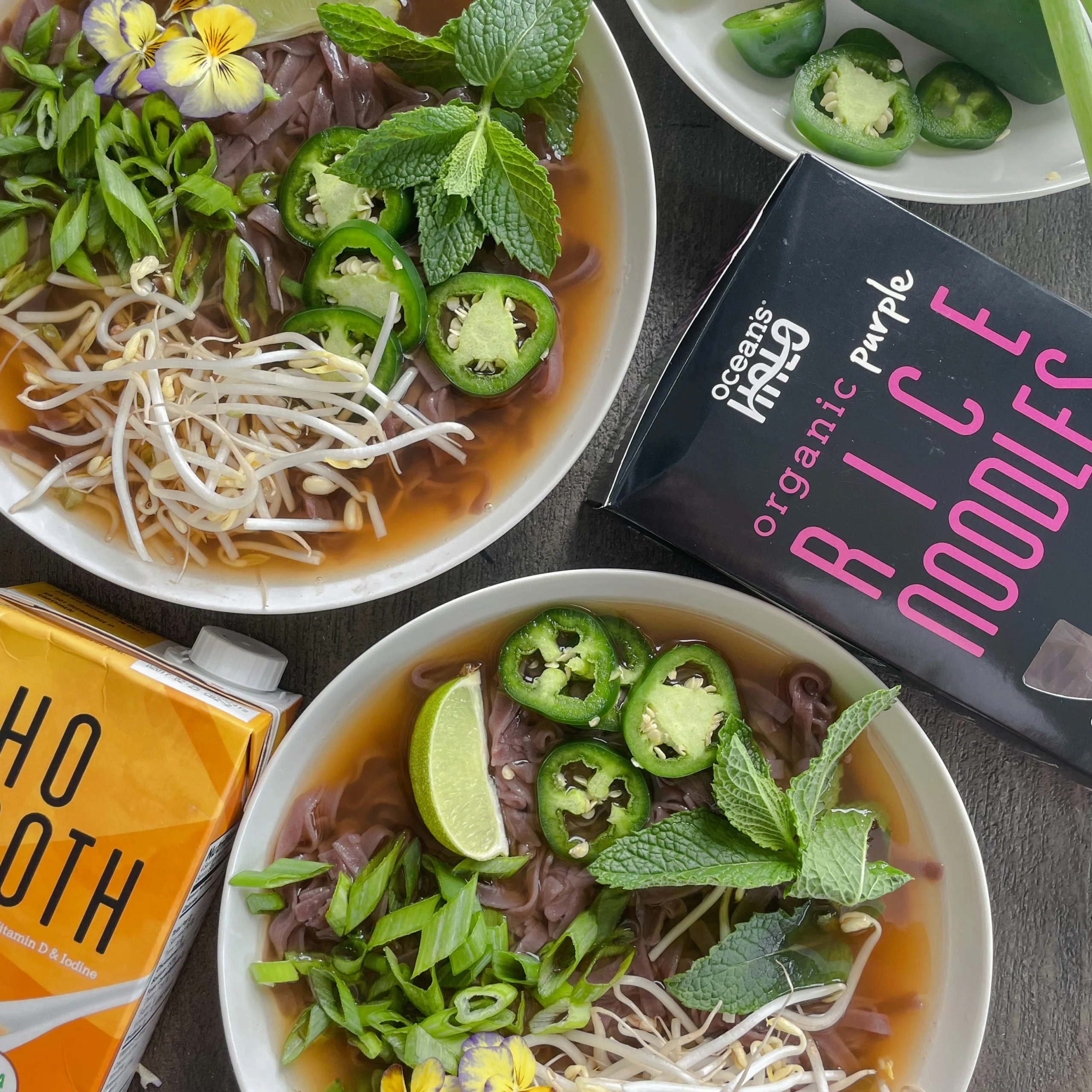 Two bowls of purple pho noodle soup with toppings, next to Ocean's Halo Pho Broth and Purple Rice Noodles packaging