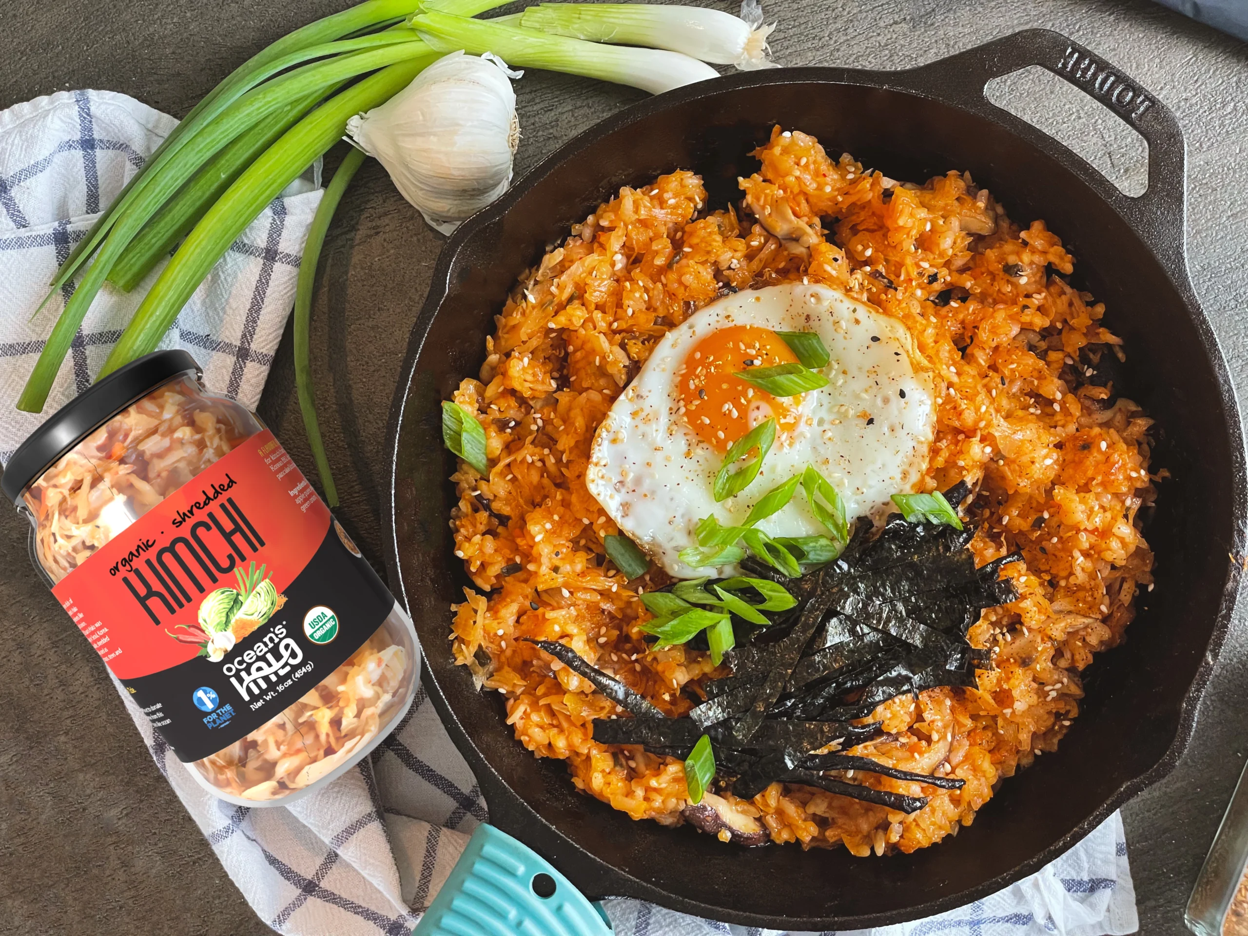 Kimchi fried rice topped with a sunny-side-up egg, sliced green onions, and seaweed strips in a black cast-iron skillet next to a jar of Ocean's Halo kimchi and a box of sticky rice.