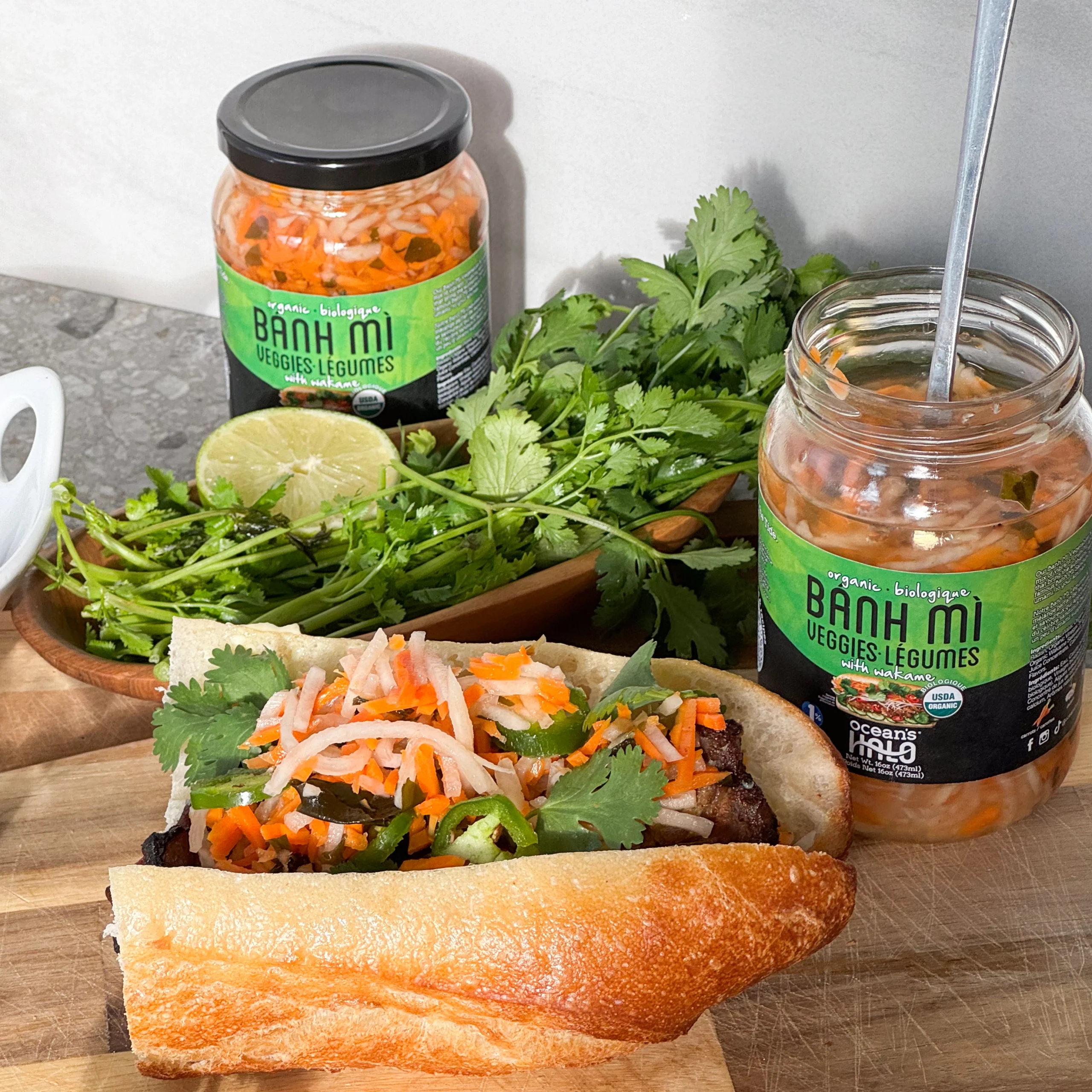 A fresh b?nh m? sandwich stuffed with pickled veggies, cilantro, and meat next to jars of B?nh M? Veggies