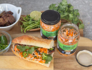 A banh mi sandwich next to jars of Ocean's Halo Banh Mi pickled vegetables.