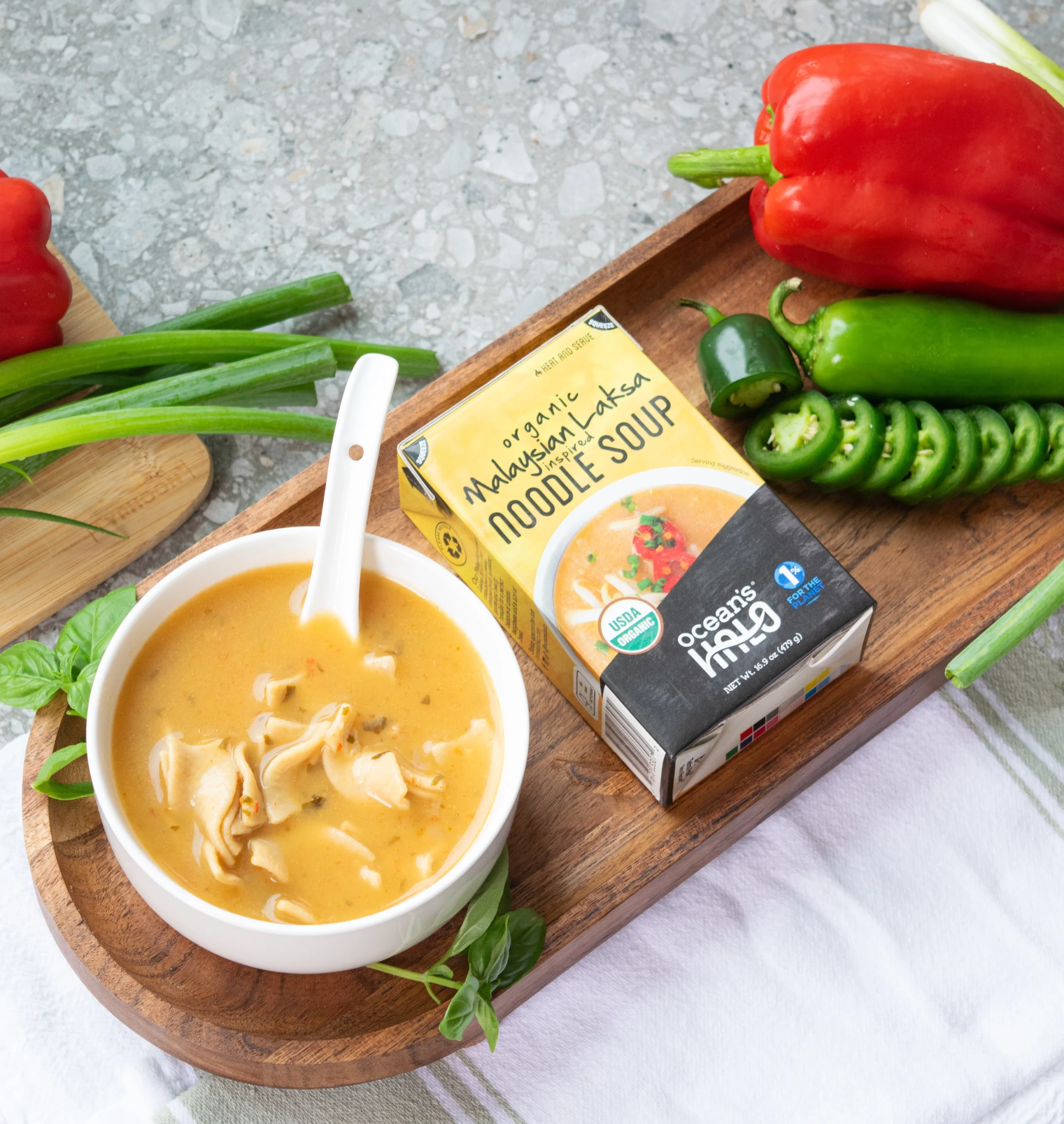 A bowl of noodle soup next to a carton of Ocean's Halo Organic Malaysian Laksa Inspired Noodle Soup on a wooden tray, surrounded by fresh ingredients like bell peppers, jalape?os, and green onions.