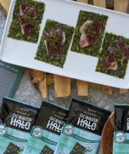 A white platter holds four seaweed snacks topped with iberico ham and caviar, surrounded by Ocean's Halo snack packs.