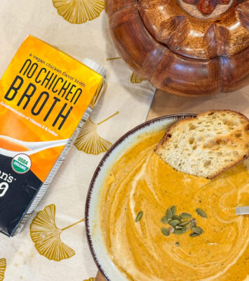 A bowl of creamy pumpkin soup with toast and seeds sits beside a carton of vegan "No Chicken Broth" on a fall placemat.