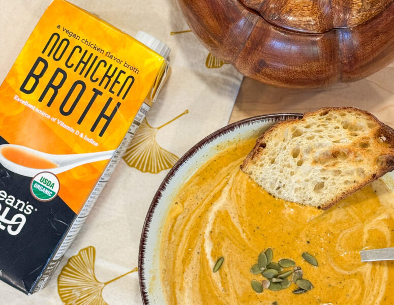 A bowl of creamy pumpkin soup with toast and seeds sits beside a carton of vegan "No Chicken Broth" on a fall placemat.
