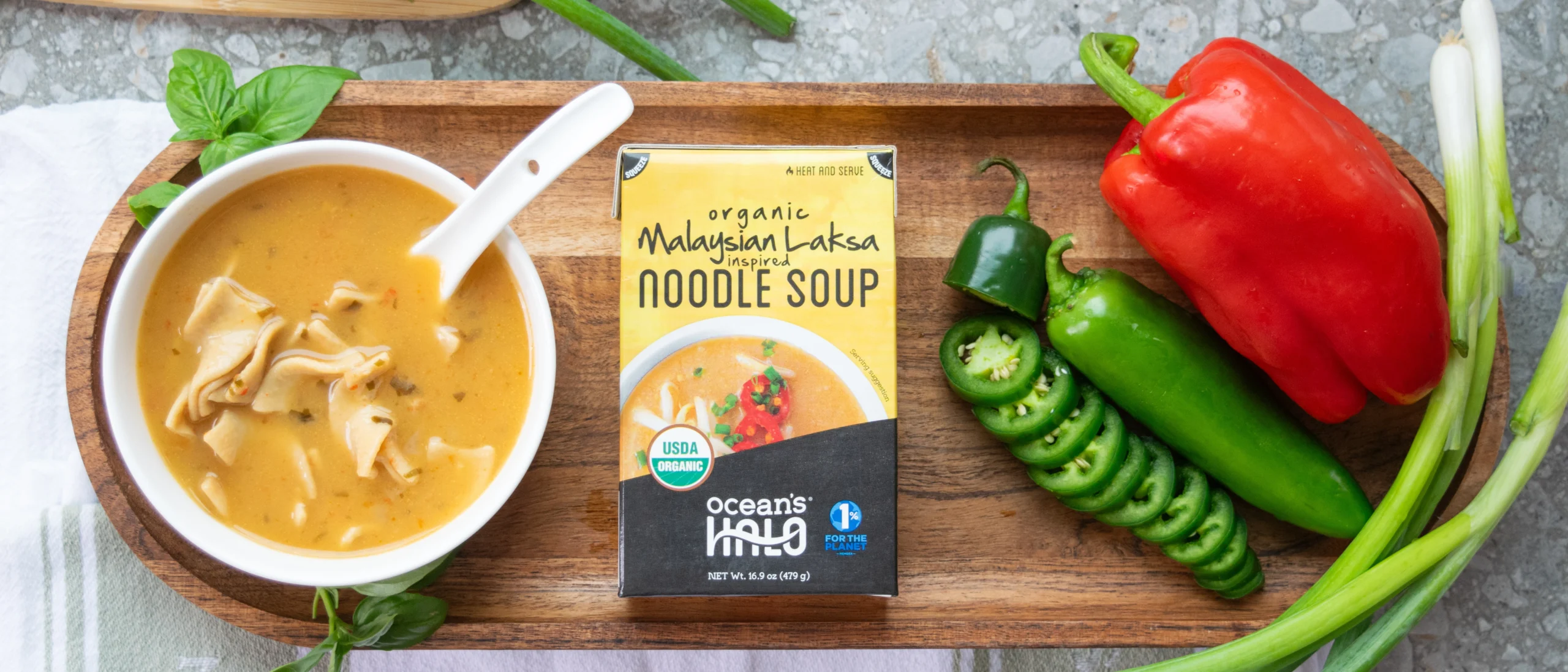 Laksa noodle soup and carton on a wooden tray with fresh red pepper, jalapeños, and green onions.
